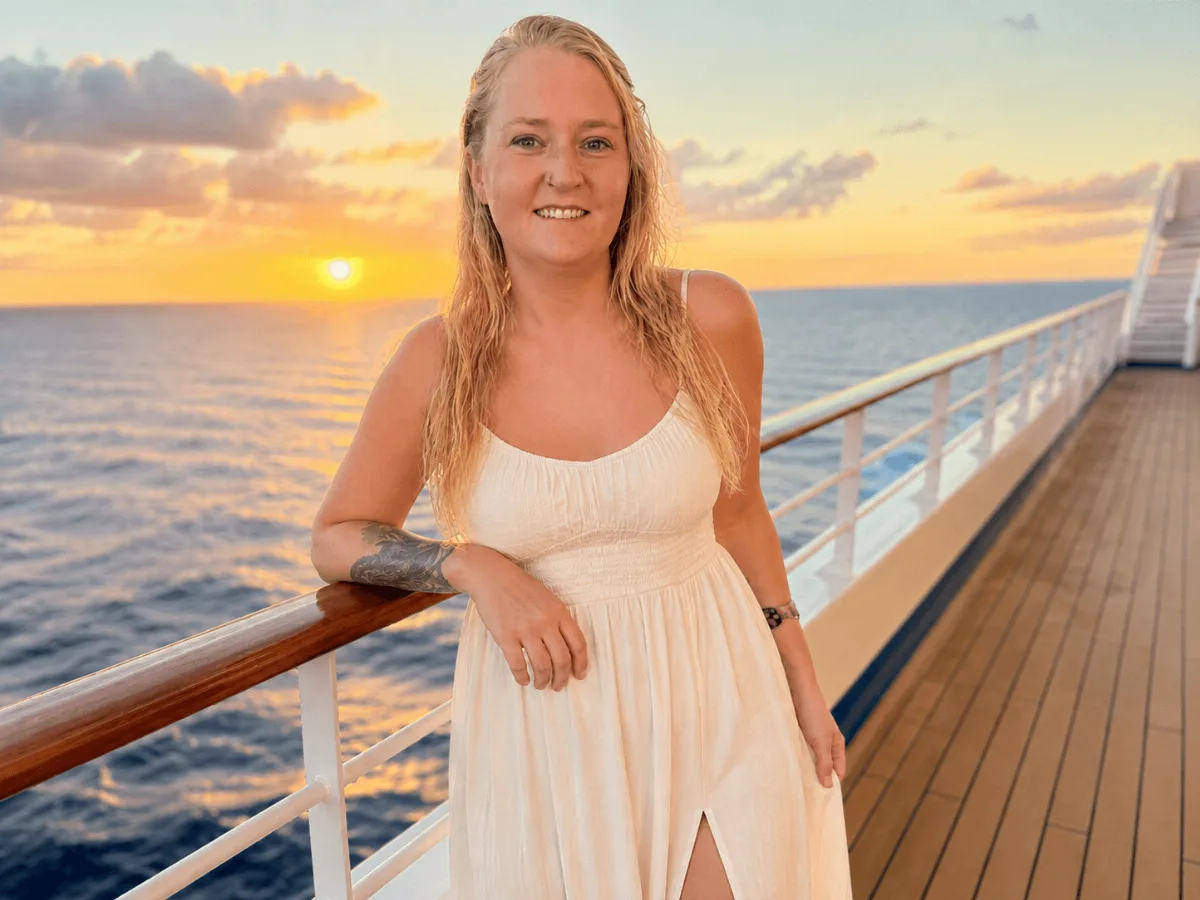 A woman standing alone at a cruise ship railing at sunset, smiling